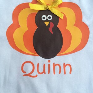 May include: White t-shirt featuring a cartoon turkey design with orange, yellow, and brown feathers. The turkey has a yellow bow and the name "Quinn" in orange script below the turkey.