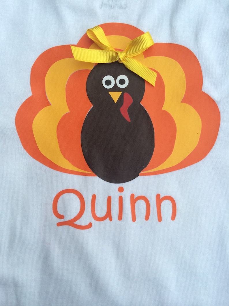 May include: White t-shirt featuring a cartoon turkey design with orange, yellow, and brown feathers. The turkey has a yellow bow and the name "Quinn" in orange script below the turkey.