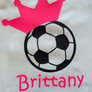 May include: White t-shirt featuring a soccer ball design with a pink crown above it. The name "Brittany" is printed in pink below the soccer ball. The soccer ball is black and white.