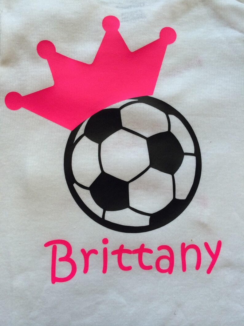May include: White t-shirt featuring a soccer ball design with a pink crown above it. The name "Brittany" is printed in pink below the soccer ball. The soccer ball is black and white.
