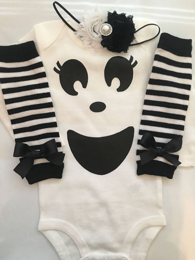 May include: A white baby bodysuit with a large black ghost face design, paired with black and white striped leg warmers and a matching headband. The leg warmers have black bows at the ankles.