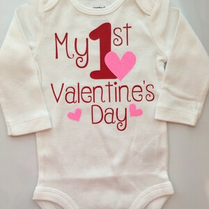 K&ouml;nnte beinhalten: Wei&szlig;er Baby-Body mit langen &Auml;rmeln. Der Body tr&auml;gt den roten Text "My 1st Valentine's Day", eine gro&szlig;e rote "1" und ein pinkfarbenes Glitzerherz. Zwei kleinere rosa Herzen sind ebenfalls abgebildet.