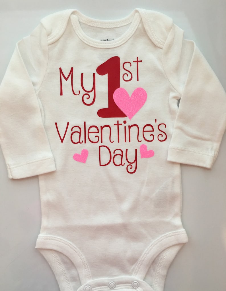 K&ouml;nnte beinhalten: Wei&szlig;er Baby-Body mit langen &Auml;rmeln. Der Body tr&auml;gt den roten Text "My 1st Valentine's Day", eine gro&szlig;e rote "1" und ein pinkfarbenes Glitzerherz. Zwei kleinere rosa Herzen sind ebenfalls abgebildet.