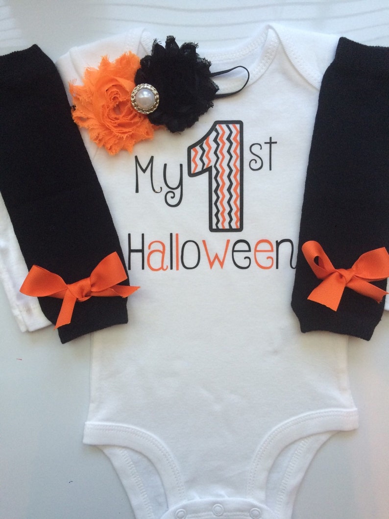 May include: A white baby bodysuit with the text "My 1st Halloween" in black and orange. The number "1" has an orange and black zig-zag design. Black leg warmers with orange bows and a floral headband complete the outfit.
