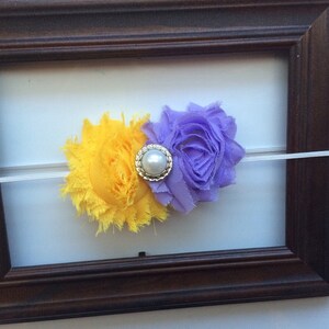 May include: A yellow and purple floral headband with a pearl and silver embellishment. The headband is made of soft fabric and is attached to a thin, white elastic band. The flowers are set against a white background.