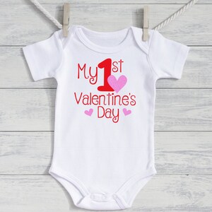 K&ouml;nnte beinhalten: Wei&szlig;er Baby-Body mit dem Text "My 1st Valentine's Day" in roter Schrift. Ein gro&szlig;es rosa Herz und zwei kleinere rosa Herzen akzentuieren den Text. Der Body h&auml;ngt an einer W&auml;scheleine.