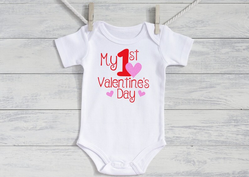 K&ouml;nnte beinhalten: Wei&szlig;er Baby-Body mit dem Text "My 1st Valentine's Day" in roter Schrift. Ein gro&szlig;es rosa Herz und zwei kleinere rosa Herzen akzentuieren den Text. Der Body h&auml;ngt an einer W&auml;scheleine.