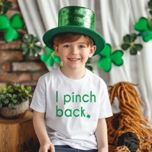 May include: A young person wearing a white t-shirt with the text "I pinch back" in green, and a green top hat. The background includes shamrock decorations, suggesting a St. Patrick's Day theme.