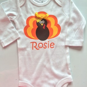 May include: White baby bodysuit with a turkey graphic in shades of orange, yellow, and brown. The turkey has a yellow bow and the name "Rosie" in orange script. The bodysuit is size 3.