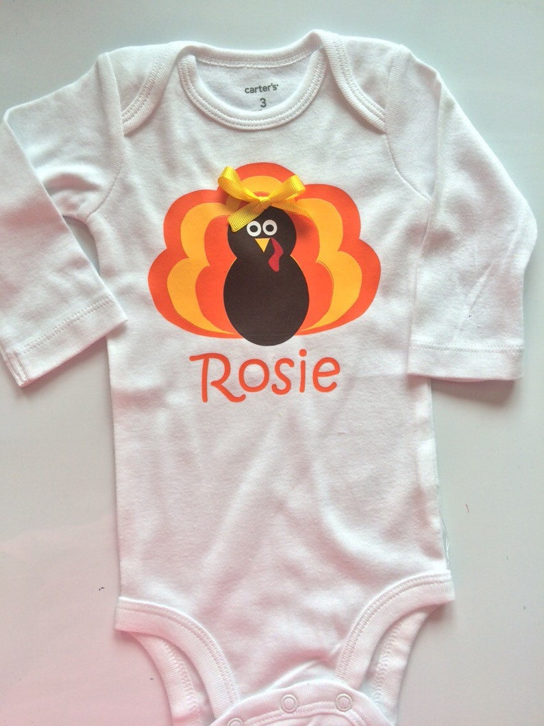 May include: White baby bodysuit with a turkey graphic in shades of orange, yellow, and brown. The turkey has a yellow bow and the name "Rosie" in orange script. The bodysuit is size 3.