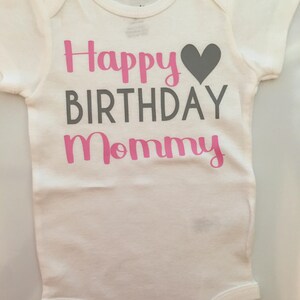 May include: White baby onesie with the text "Happy Birthday Mommy" in pink and gray lettering, with a gray heart graphic. The onesie has short sleeves and a simple design.