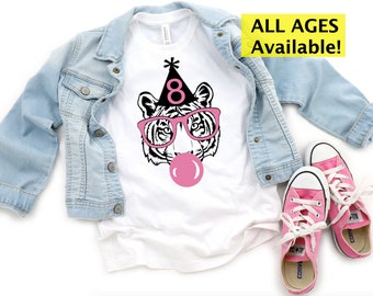 May include: White t-shirt with a black and white tiger face wearing pink glasses and a party hat with the number 8. A pink bubble gum bubble is in front of the tiger. The shirt is paired with a light blue denim jacket and pink sneakers. Text on the image says "ALL AGES Available!"
