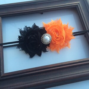 May include: A black and orange floral headband with a pearl-centered embellishment. The headband features two fabric flowers, one black and one orange, attached to a thin black elastic band. The design is set against a white background.