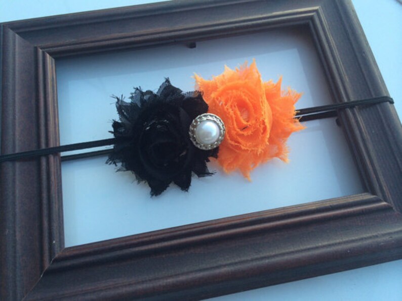 May include: A black and orange floral headband with a pearl-centered embellishment. The headband features two fabric flowers, one black and one orange, attached to a thin black elastic band. The design is set against a white background.