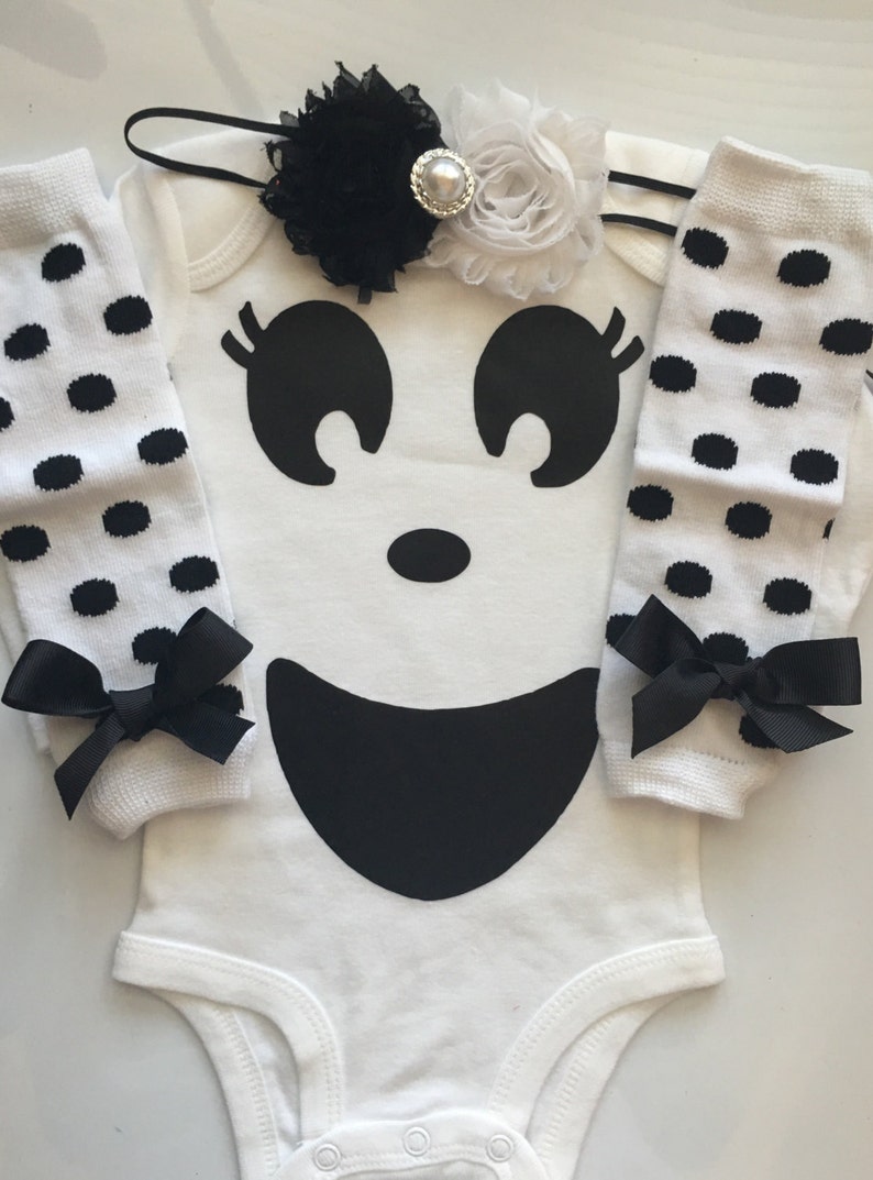 May include: A white baby bodysuit with a large black ghost face design, paired with matching white leg warmers with black polka dots and black bows. A black and white floral headband with a pearl center completes the set.