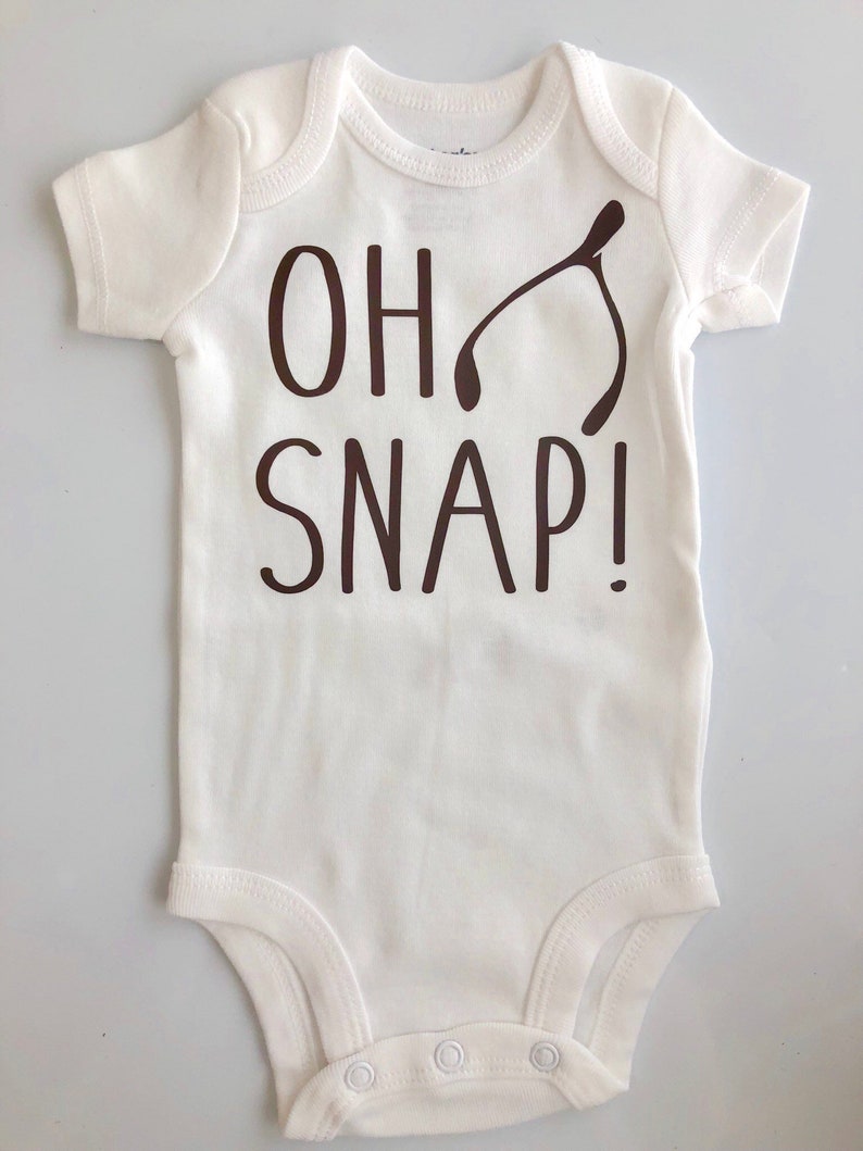 May include: A white baby onesie with short sleeves and a round neckline. The words "OH SNAP!" are printed in dark brown, along with a wishbone graphic. The onesie has snap closures at the bottom.