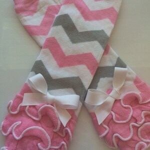 May include: Baby leg warmers featuring a pink, white, and grey zigzag pattern. The cuffs are pink with a frilled design and white trim, finished with white bows.