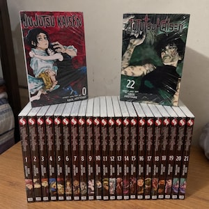 Könnte beinhalten: Eine Sammlung von Jujutsu Kaisen Manga-Bänden von Gege Akutami, mit Band 0 und Band 22 aufrecht stehend, sowie einer Reihe von Bänden nummeriert von 1 bis 21, die horizontal darunter angeordnet sind, alle mit illustrierten Buchrücken und Covern.
