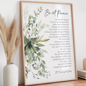 Può includere: Una stampa incorniciata con le parole "Be at Peace" in una scrittura elegante. La stampa presenta un disegno ad acquerello di foglie e fogliame verdi. Il testo è una citazione di San Francesco di Sales.