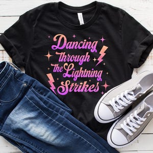 Könnte beinhalten: Ein schwarzes T-Shirt mit dem Text "Dancing Through the Lightning Strikes" in einem Farbverlauf von Pink und Lila. Dazu eine blaue Jeans und graue Leinenschuhe mit weißen Schnürsenkeln.