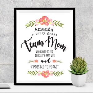 May include: A framed print with the words "Amanda a truly great Team Mom" in black script. The print is decorated with watercolor floral accents in pink and green. A small succulent in a white pot sits to the right.