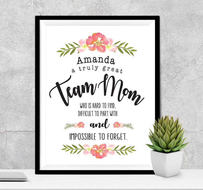 May include: A framed print with the words "Amanda a truly great Team Mom" in black script. The print is decorated with watercolor floral accents in pink and green. A small succulent in a white pot sits to the right.