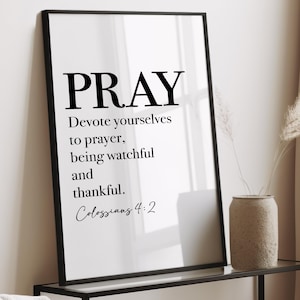 Pode incluir: Arte de parede emoldurada em preto com a palavra "PRAY" em letras pretas e em negrito. Abaixo, o texto diz "Devote yourselves to prayer, being watchful and thankful. Colossians 4:2". A arte é exibida em uma prateleira.