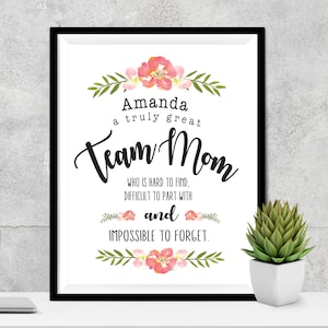 May include: A framed print with the words "Amanda a truly great Team Mom" in black script. The print is decorated with watercolor floral accents in pink and green. A small succulent in a white pot sits to the right.