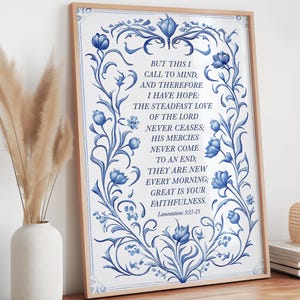 May include: A framed print featuring a quote from Lamentations 3:21-23, with a blue floral design on a white background. The text is centered within a decorative border, and the frame is a light wood color. The print is displayed on a wooden surface.