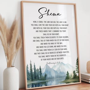 Può includere: Una stampa incorniciata con la parola "Shema" in corsivo. Il testo di Deuteronomio 6:4-9 è visualizzato sotto un paesaggio ad acquerello di montagne, alberi e un lago. La cornice è di colore legno chiaro.