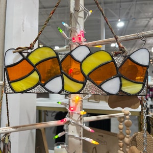May include: A stained glass window hanging ornament featuring a row of candy corn in orange, yellow, and white. The ornament is hanging from a white wooden stand with string lights.