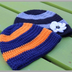 Op de afbeelding: Twee gebreide mutsen, een met blauwe en oranje strepen en de andere met grijze en zwarte strepen. De muts met de blauwe en oranje strepen heeft een witte gebreide bloem bovenop.