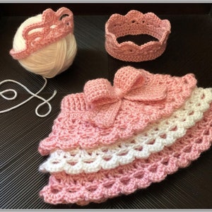 May include: Pink crocheted crown and skirt set with a white yarn ball. The skirt has a white and pink layered design with a bow on the front.