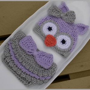 May include: A crocheted baby owl hat and diaper set in shades of purple and gray. The hat features a large bow and two large eyes with black pupils. The diaper has a purple bow and a gray and purple striped pattern.