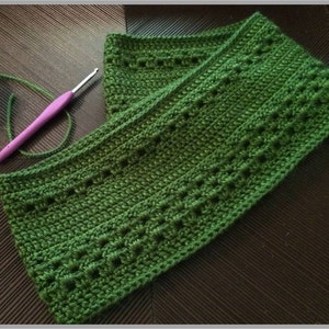 May include: A handmade, green crochet scarf with a textured pattern. A purple and silver crochet hook rests on the scarf, with green yarn extending from the hook. The scarf is folded, showcasing the intricate stitch work.
