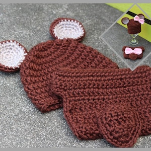 May include: A brown crocheted baby hat and matching diaper cover with white and pink accents. The hat has two floppy ears and the diaper cover has a bow.