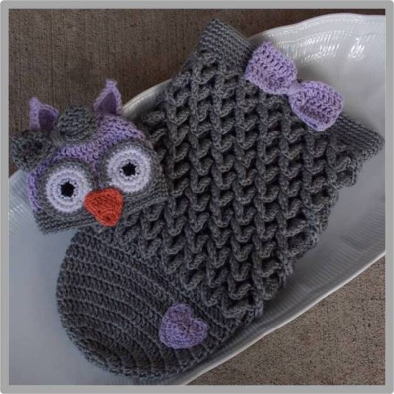 May include: A gray crocheted baby sleeping bag with an attached purple and gray owl hat. The sleeping bag has a purple heart on the bottom and a purple bow on the top.