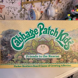 May include: A vintage Cabbage Patch Kids board game box with a yellow background and green text. The text reads "Cabbage Patch Kids Friends to the Rescue" and "Parker Brothers Board Game of Growing Affection". The box features an illustration of a Cabbage Patch Kid doll with green leaves and a smiling face.