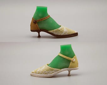 May include: Two pairs of miniature doll shoes. One pair is tan and brown with a lace overlay, and the other is yellow with a lace overlay. Both have a strap and a small heel. The shoes are displayed on green foot forms.