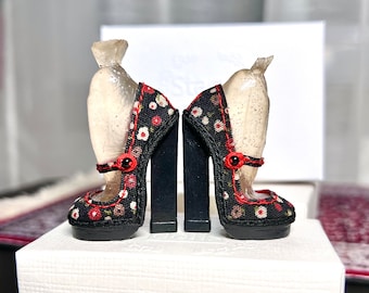 May include: A pair of black high-heeled shoes with a floral print and red accents. The shoes have a chunky heel and a closed toe.