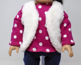 May include: A doll wearing a magenta long-sleeved shirt with white polka dots, a white fluffy vest, navy blue pants, and white fluffy boots. The doll has brown hair and is standing on a white stand.