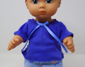 May include: A doll wearing a blue top with a bow, light blue pants with a bird pattern, a pink lace headband, and white shoes. The doll has blue eyes and a fair complexion. The doll is a toy.
