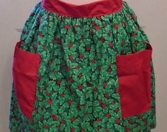 May include: A Christmas-themed apron with a green holly and red berry pattern. The apron has two large red pockets and a red waistband. The apron is designed to be worn around the waist and is made of fabric.