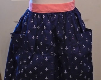 May include: A navy blue apron with white anchor patterns and two front pockets. The apron has a coral pink waistband. The apron is displayed on a mannequin. The apron is a nautical theme.