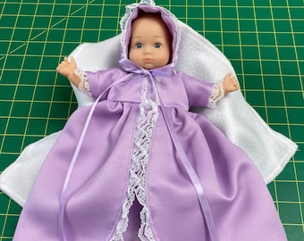 May include: A baby doll wearing a lavender satin christening gown with white lace trim and a matching bonnet. The gown has a flowing skirt and is tied with a lavender ribbon. The doll is lying on a white blanket.