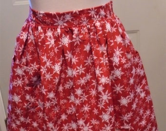 May include: A red apron with a white snowflake pattern. The apron has two pockets and a gathered waist. The apron is made of a lightweight fabric.