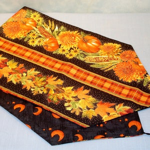 Può includere: Runner da tavolo reversibile a tema autunnale con motivi di zucche, foglie e fiori su sfondo scuro. Un lato presenta un motivo di zucche e fiori, l'altro ha lune e stelle arancioni. Nappe nere alle estremità.
