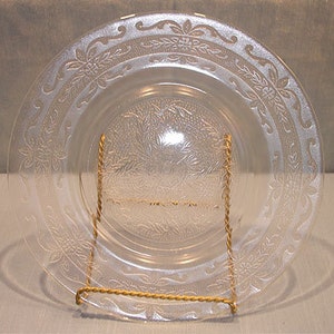 Puede incluir: Un plato de vidrio transparente con un diseño floral y de hojas en relieve. El plato es redondo y tiene un borde decorativo. El plato se exhibe en un soporte de metal dorado.