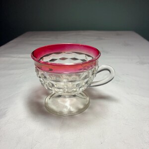 May include: A clear glass cup featuring a faceted diamond pattern on the exterior, a pedestal base, and a curved handle. The upper rim of the cup is accented with a vibrant, solid pink band that encircles the entire top edge of the vessel.