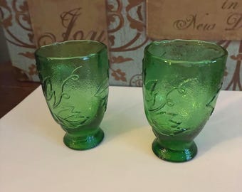 May include: Two matching green glass tumblers featuring a textured surface with an embossed leaf and vine pattern. Each tumbler has a flared rim, a tapered body, and a circular pedestal base, creating a consistent look for both drinking vessels.
